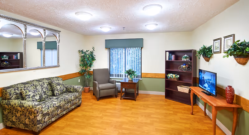 Community living room with a patterned sofa, armchair, side tables, bookshelf, TV on a console, plants and a large wall mirror.