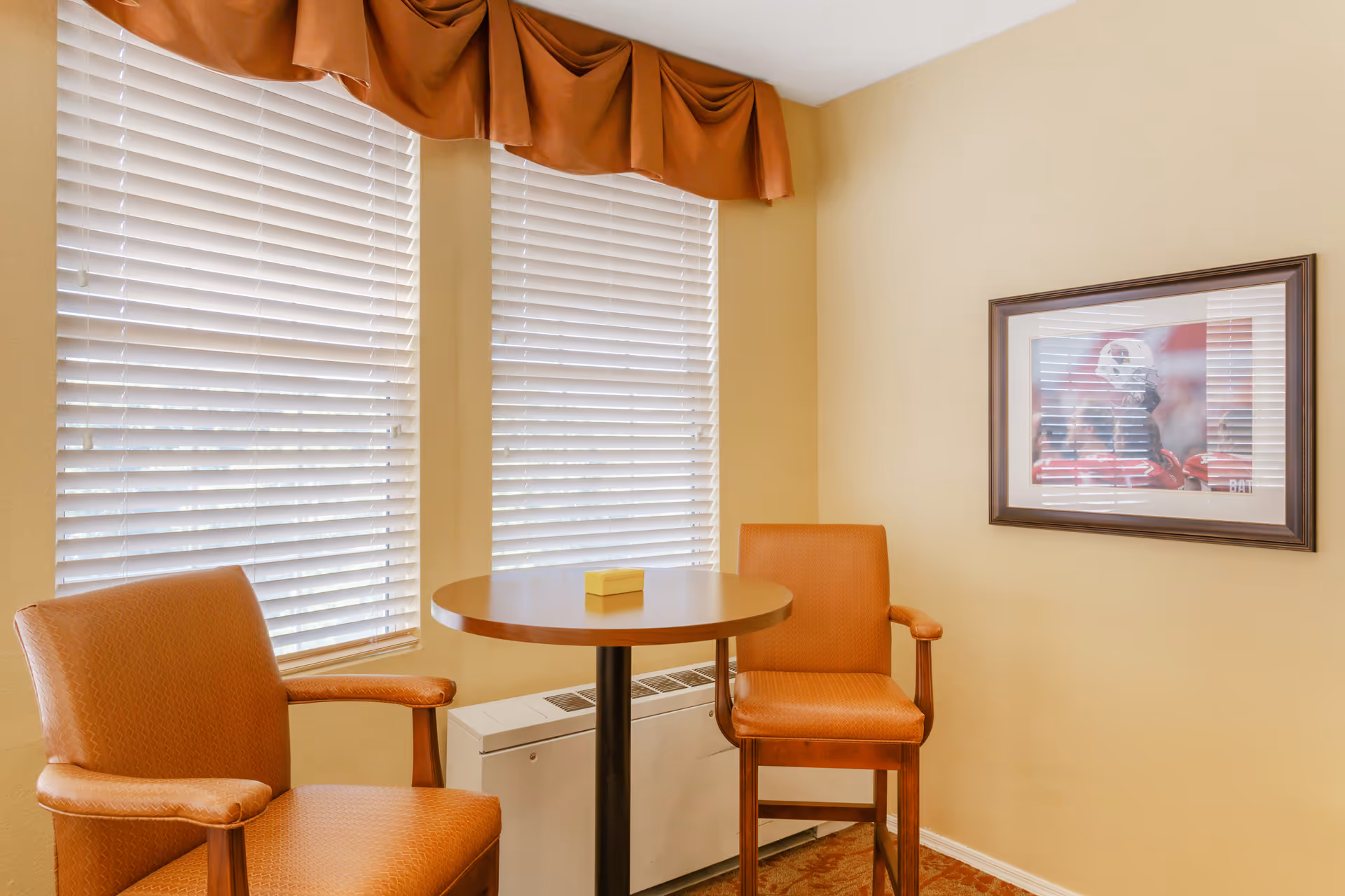 A small seating area with two orange upholstered chairs and a round wooden table between them. The background features two windows with white blinds and an orange valance. A framed picture hangs on the beige wall to the right.