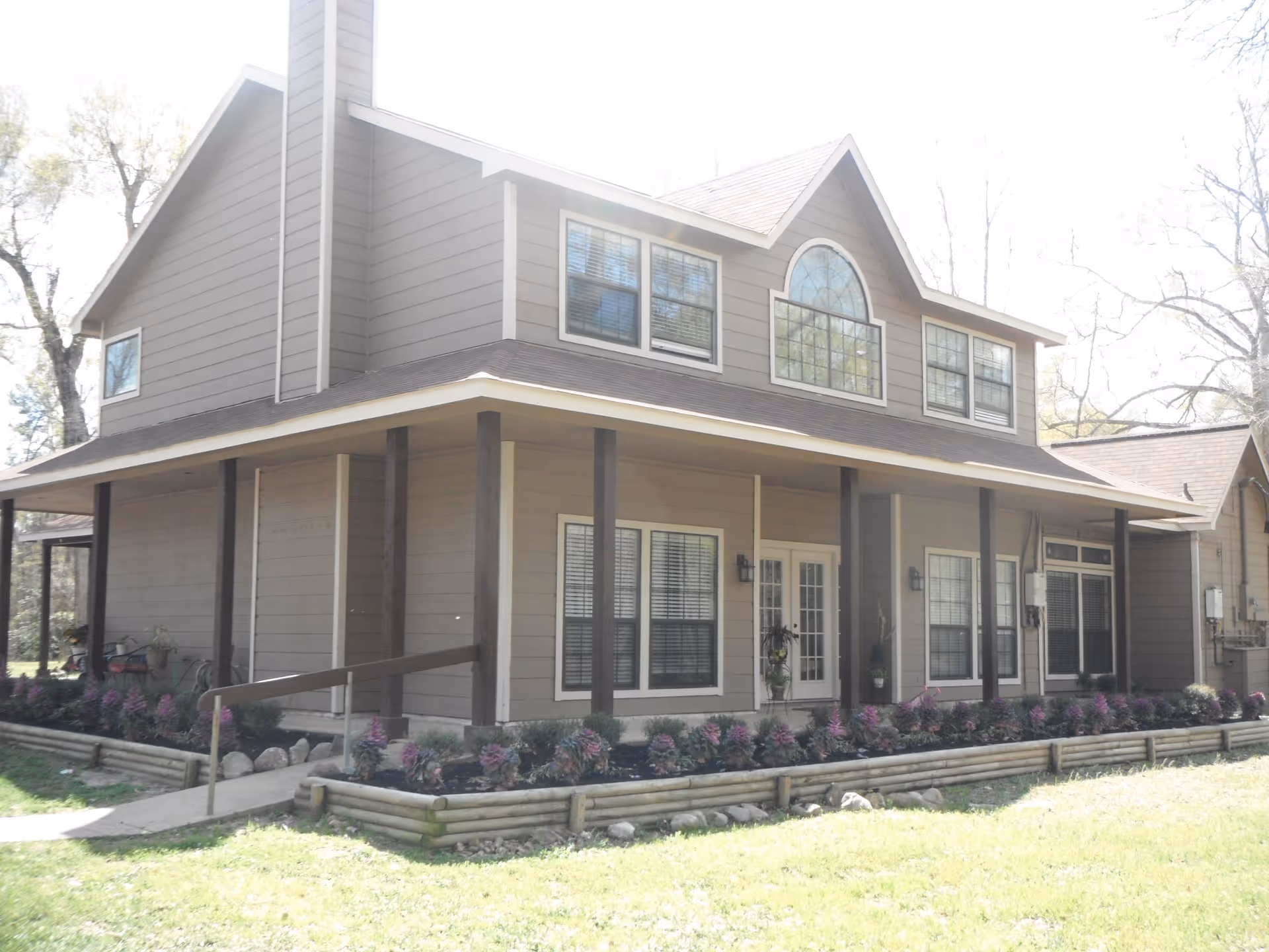 Exterior view of a two-story residential building with beige siding and dark brown trim. The building features multiple windows, a large arched window on the upper floor, and a covered porch supported by dark brown columns. There is a small garden bed with purple and green plants bordered by wooden logs in front of the porch, and a concrete walkway with a handrail leading to the entrance.