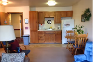 Small apartment-style interior showing a kitchenette with wooden cabinets and fridge, a dining table with chairs, and living room seating.