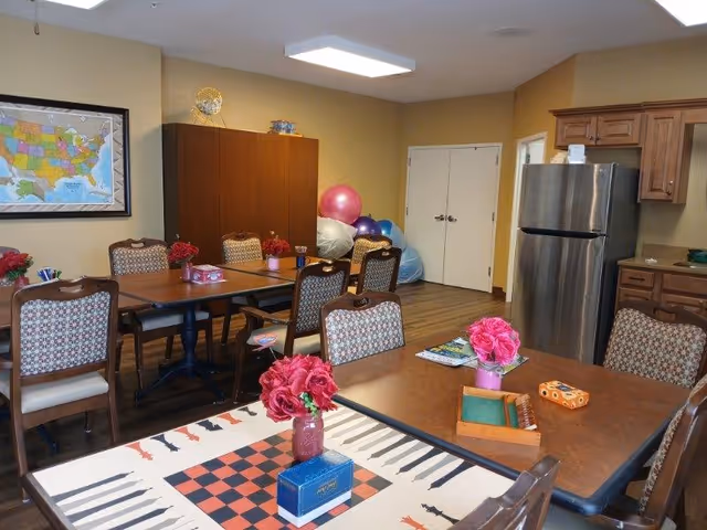 A communal room with several tables and chairs arranged for group activities. The tables have decorative flowers and board games on them. In the background, there is a stainless steel refrigerator, wooden cabinets, and a set of double doors. Exercise balls and bean bags are stacked in the corner. A framed map of the United States hangs on the wall.