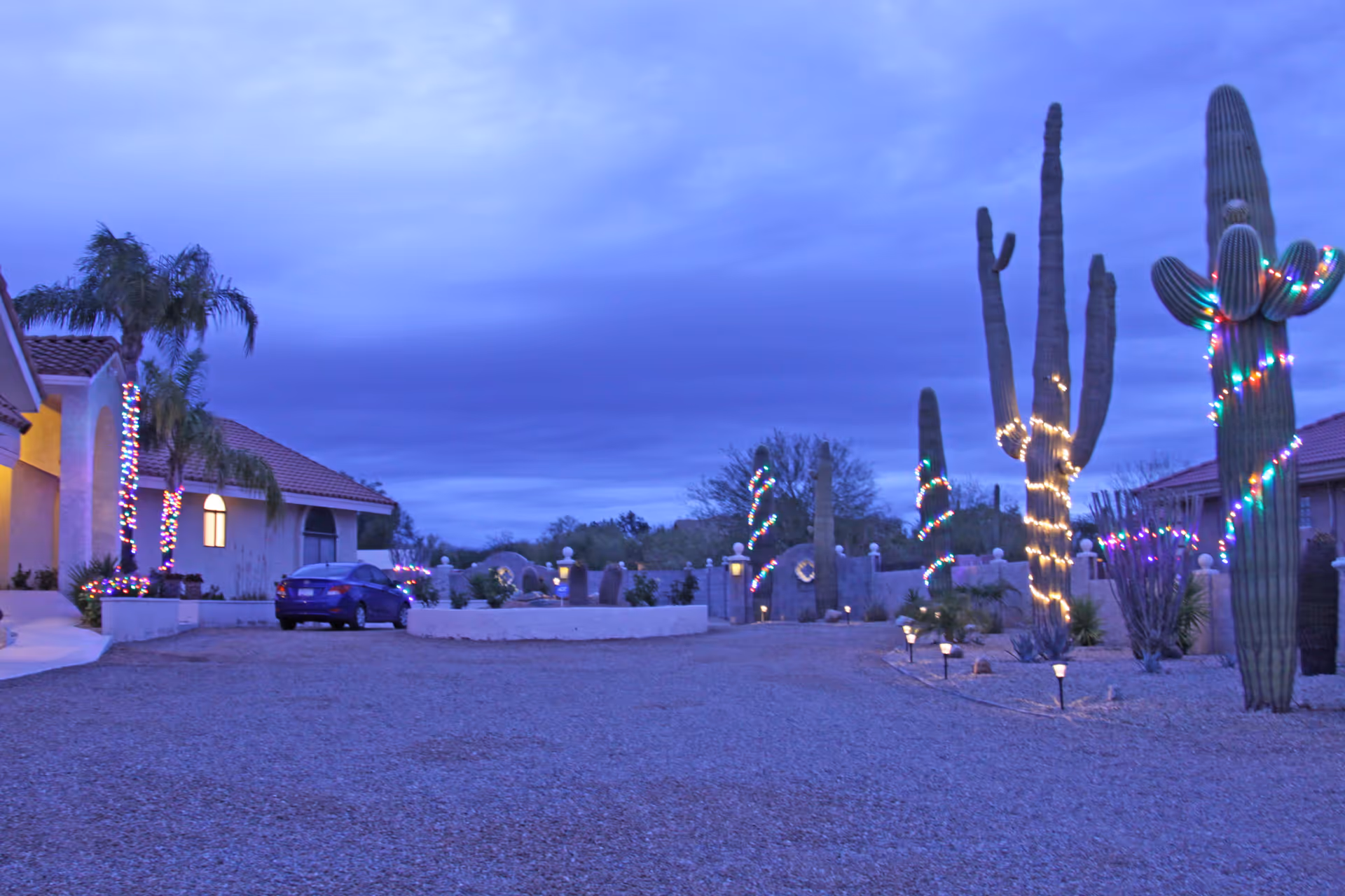 Dusk exterior of a desert-style assisted living property with cacti and palm trees wrapped in colorful lights, tiled-roof buildings, and a parked car.