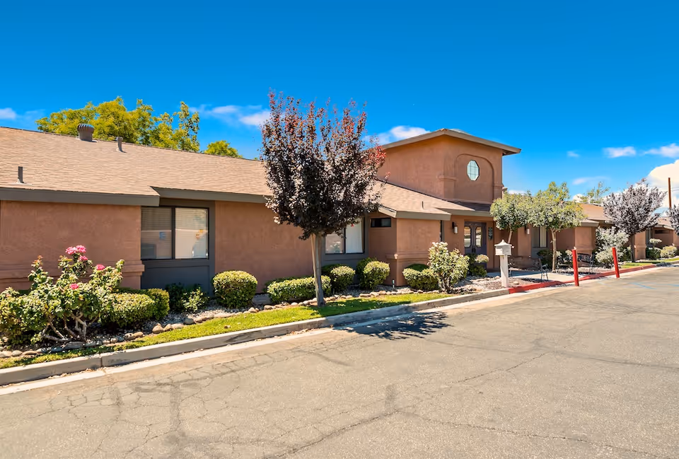 Single-story stucco memory care building with landscaped shrubs and trees along a paved driveway under a clear blue sky.