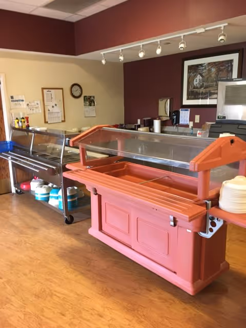 A dining service area with two food serving stations, one orange and one metallic, both with clear sneeze guards. Plates are stacked on the right side of the orange station. The background shows a counter with various items, a clock on the wall, a framed picture, and a calendar.