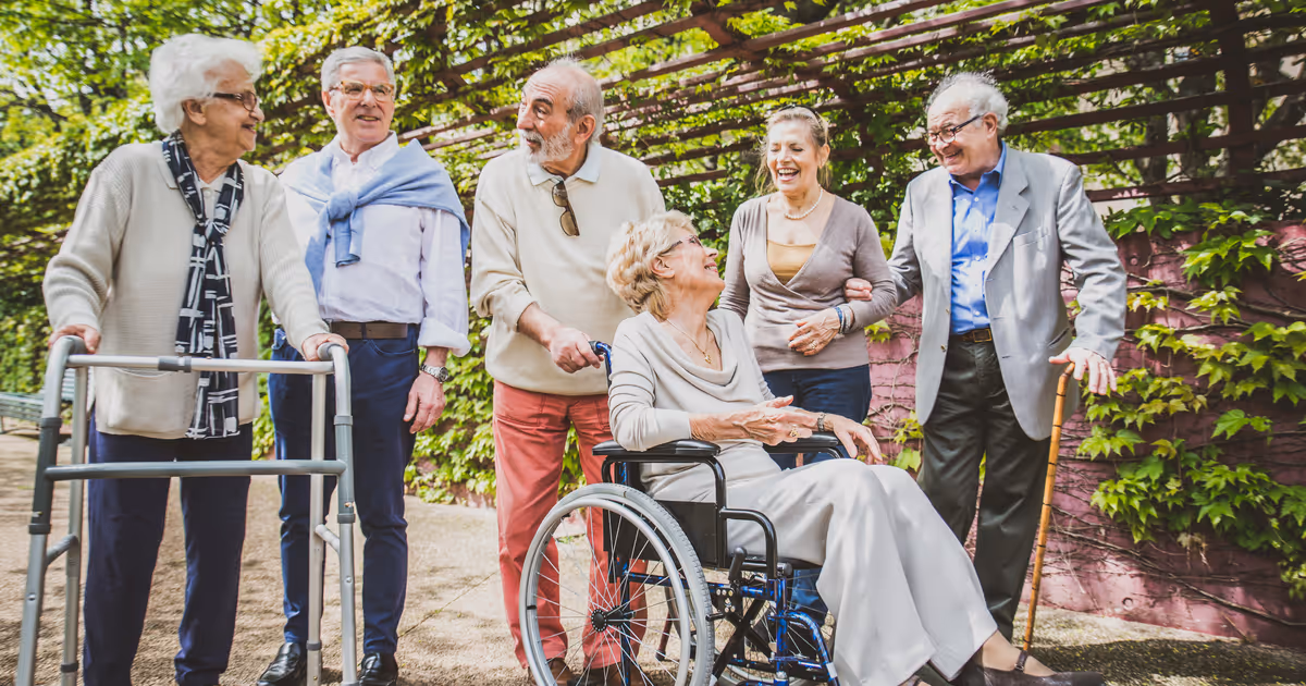 A group of six elderly people outdoors, with one woman in a wheelchair being pushed by a man. They are smiling and engaging with each other in a garden-like setting with green foliage on a wall behind them.