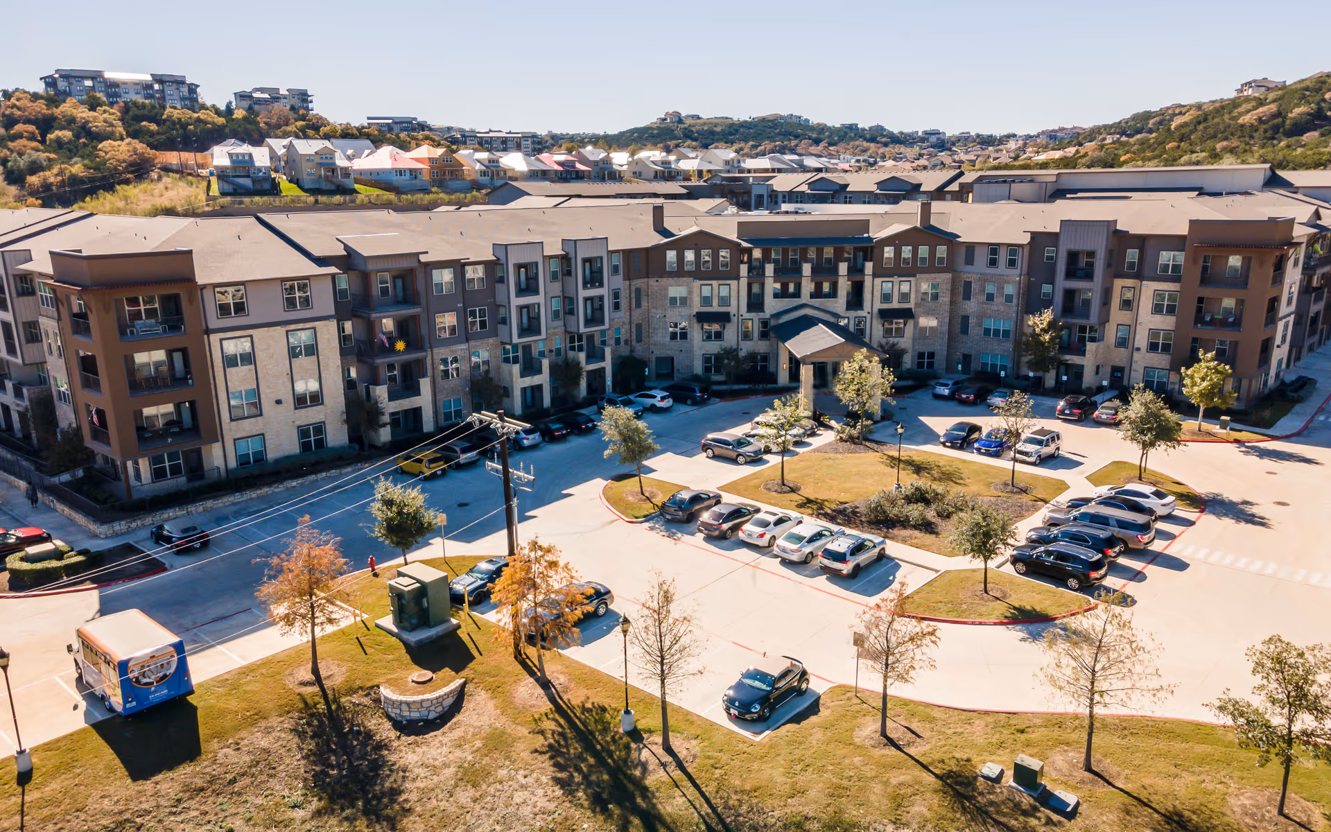 Aerial view of Discovery Village At Dominion, showing a large multi-story residential building with a parking lot in front. The building is surrounded by trees and greenery, with hills and houses visible in the background under a clear sky.