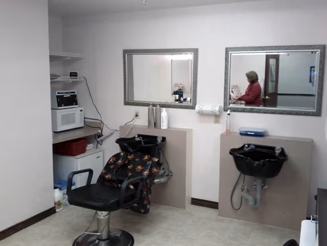 Interior of a small salon area with two black hair washing sinks mounted on beige counters, a black salon chair with a floral cape draped over it, two large mirrors on the wall, and a woman reflected in one of the mirrors. There is a microwave and other small appliances on a shelf in the corner.