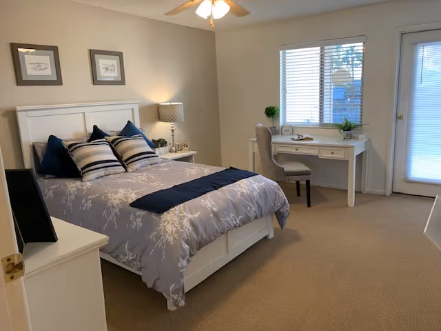 A neatly arranged bedroom with a white bed frame and a bed made with gray and white bedding, accented by several pillows. There is a white nightstand with a lamp and decorative items next to the bed. A white desk with a chair is positioned under a window with blinds, and a door with a glass panel is visible to the right. The room has beige carpeting and neutral-colored walls.