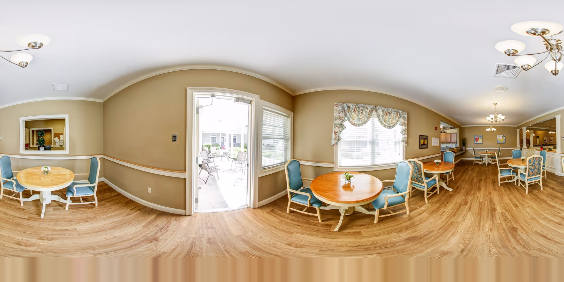 Bright communal dining room with round wooden tables and teal upholstered chairs and a door opening to an outdoor patio.