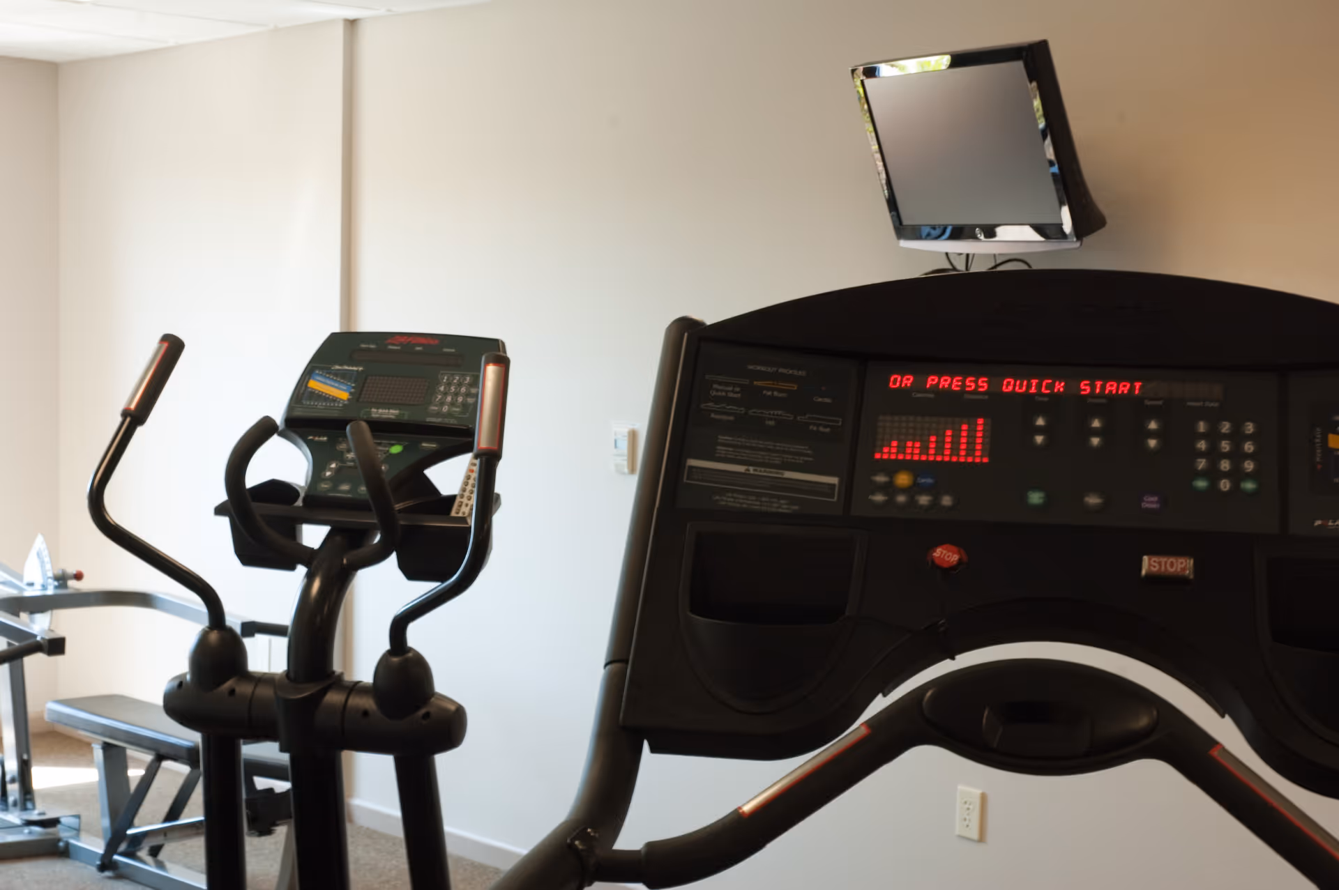 Treadmill and elliptical machines in a small exercise room with a wall-mounted TV.