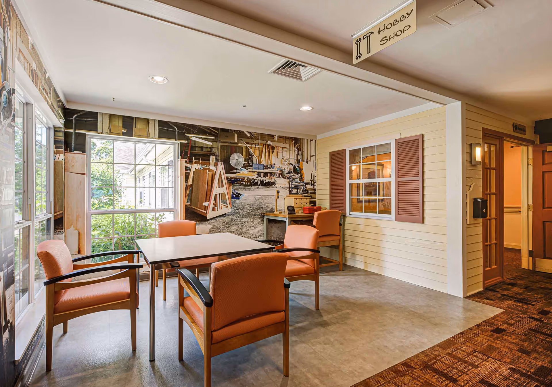 A cozy hobby shop area inside a senior living facility with a square table surrounded by four orange cushioned chairs. The room features large windows letting in natural light, a wall mural depicting a workshop, and a small desk with another chair. A sign hanging from the ceiling reads 'Hobby Shop'.