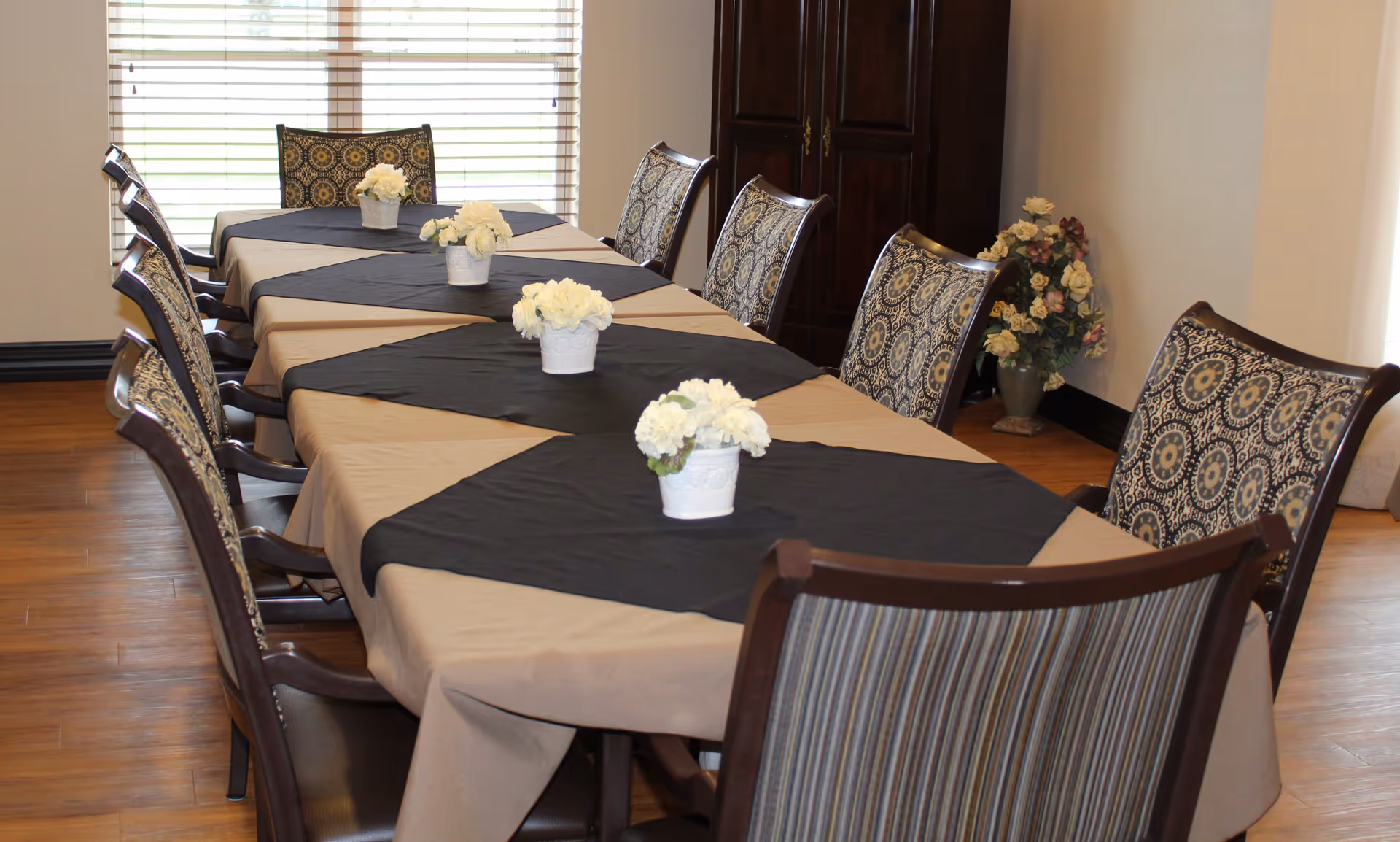 A dining room with a long rectangular table covered with a beige tablecloth and black table runners. The table is surrounded by wooden chairs with patterned upholstery. Small white flower arrangements in white pots are placed along the center of the table. In the background, there is a window with blinds and a dark wooden cabinet with a vase of flowers beside it.