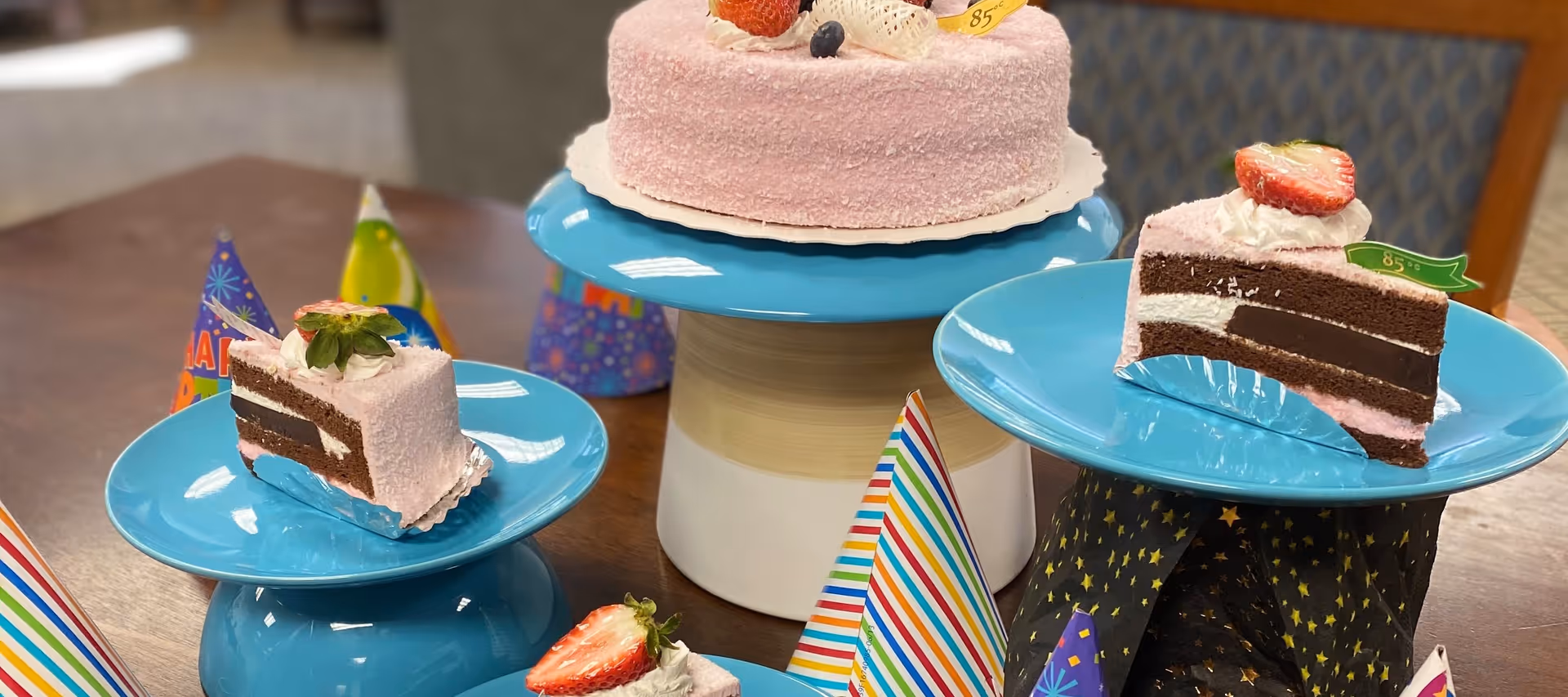 A pink frosted cake with strawberries on top displayed on a blue cake stand, with two slices of the cake on smaller blue stands around it. Colorful party hats are placed on the table around the cake and slices.