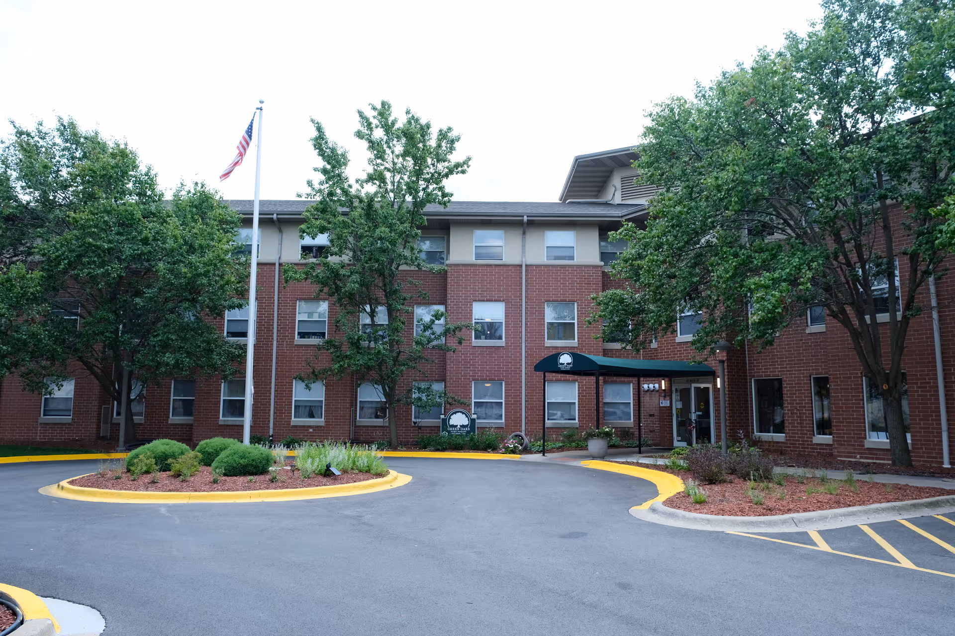Front entrance of a three-story red-brick senior living building with a circular driveway, flagpole, awning, and landscaped trees and shrubs.