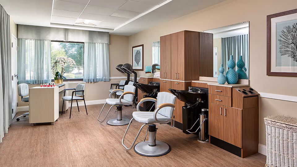 A well-lit salon room with three white salon chairs in front of wooden cabinets and black hair washing sinks. There is a manicure table with two chairs near a large window with sheer curtains. The room has light beige walls, wooden flooring, and decorative blue vases on the counter. A framed artwork hangs on the wall.