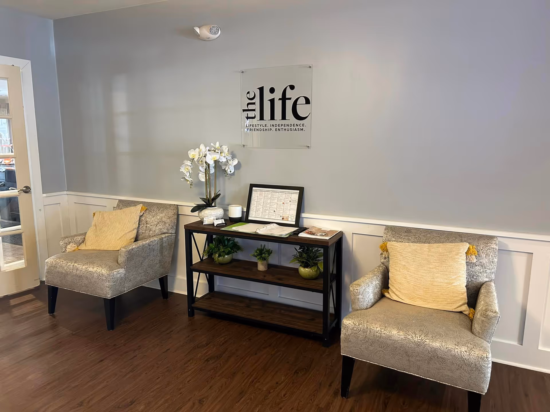 A cozy seating area with two patterned armchairs each with a yellow cushion, a wooden console table with potted plants and framed documents, and a wall sign that reads 'the life LIFESTYLE. INDEPENDENCE. FRIENDSHIP. ENTHUSIASM.'