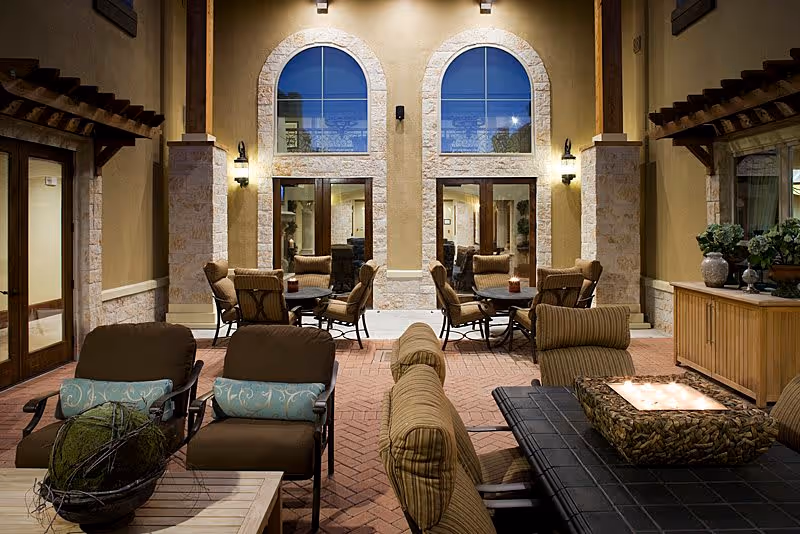 Outdoor patio area at night with cushioned chairs and tables arranged for seating. The space is surrounded by a building with large arched windows and warm lighting, creating a cozy atmosphere.