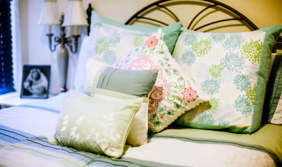 A neatly made bed with patterned and floral decorative pillows, a metal headboard, and a bedside lamp and framed photo visible.