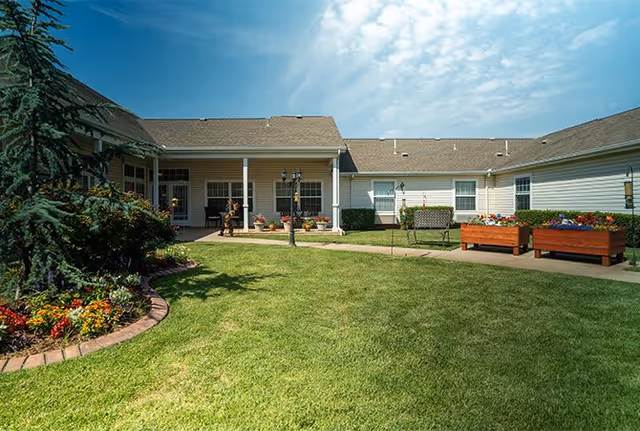 A sunny outdoor courtyard area at Morada Midwest City featuring a well-maintained green lawn, flower beds with colorful flowers, a paved walkway, a bench, and raised garden beds. The courtyard is surrounded by single-story buildings with white siding and a brown roof under a partly cloudy blue sky.