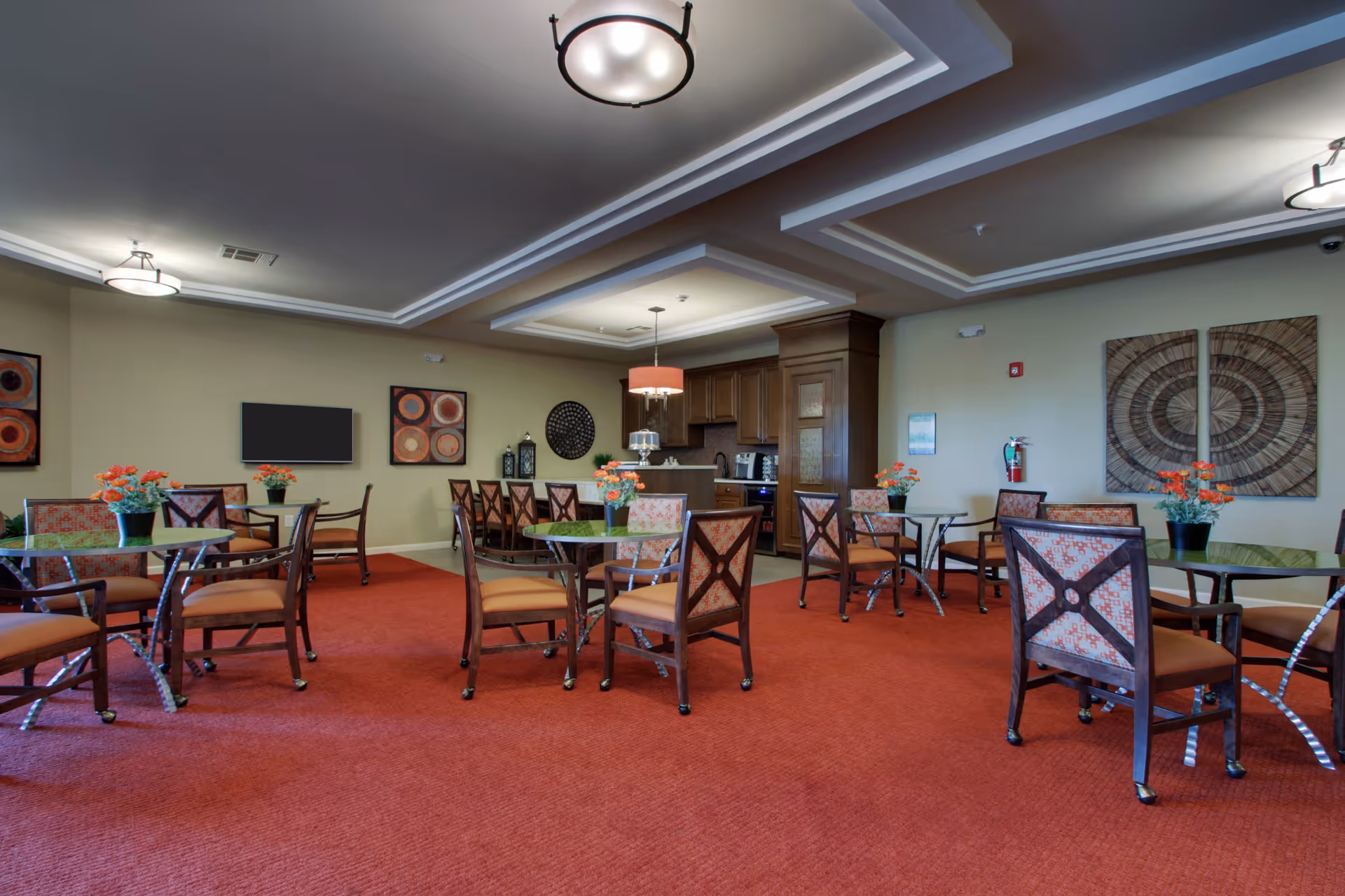 A senior living facility dining room with multiple round glass tables and wooden chairs with patterned cushions. Each table has a small flower arrangement. The room has a red carpet, beige walls with decorative artwork, and a ceiling with recessed lighting. A small kitchen area with wooden cabinets and appliances is visible in the background.