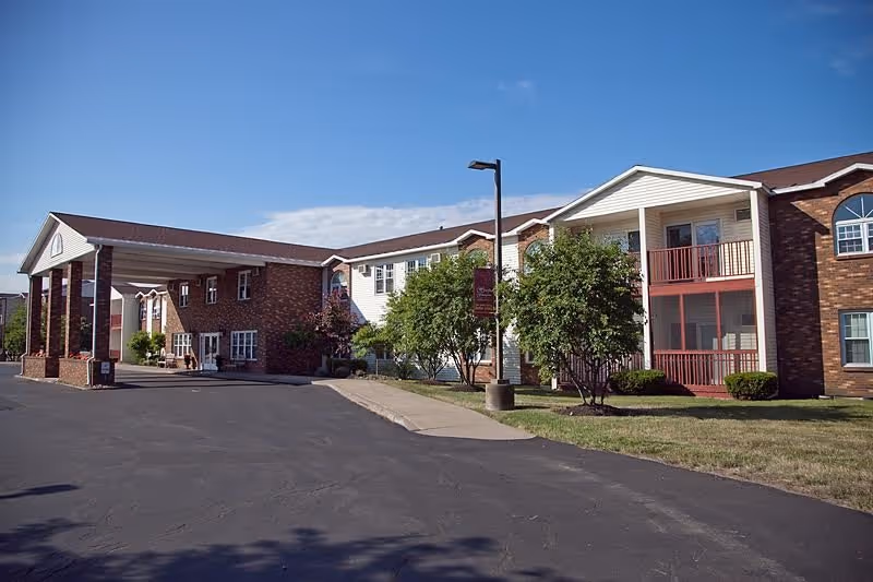 Exterior view of The Hearth at Greenpoint senior living facility showing a two-story building with brick and white siding, a covered entrance with columns, trees, and a clear blue sky.