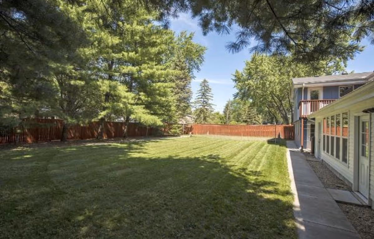 A spacious, well-maintained grassy backyard area with a concrete walkway along the right side, bordered by a wooden fence and surrounded by tall green trees under a clear blue sky. Part of a building with large windows and a balcony is visible on the right.