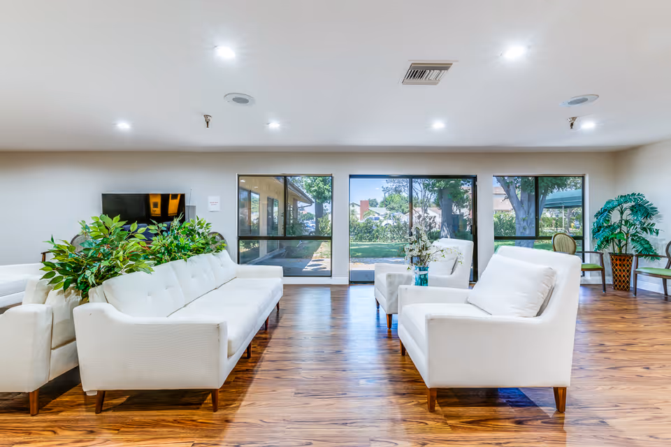Bright and spacious living room area with white sofas and armchairs arranged around a central space. Large windows provide a view of a green outdoor garden. The room has wooden flooring, recessed ceiling lights, and several potted plants adding greenery to the interior.