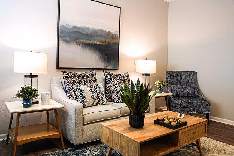 Cozy living room with a sofa, armchair, coffee table, lamps, artwork, and decorative plants.
