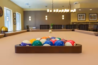 A billiards table with a full rack of colorful pool balls set up in the center of a well-lit room. The room features large windows on the left, modern light fixtures hanging from the ceiling, and framed artwork on the walls. There are chairs and tables in the background, suggesting a recreational or common area.