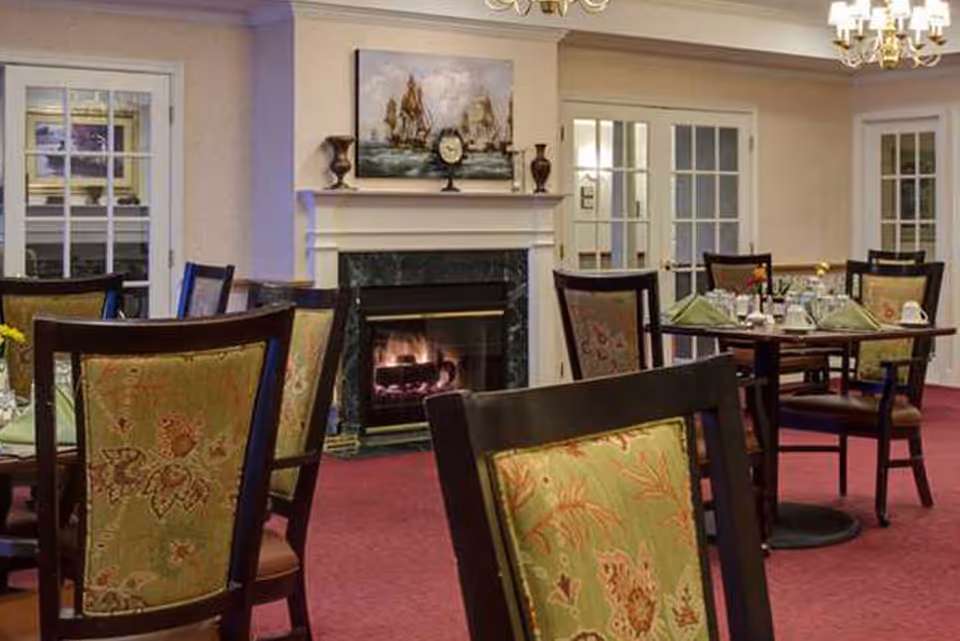 Dining room with multiple tables and chairs featuring floral upholstery, a lit fireplace with a painting and decorative items on the mantel, and glass-paneled doors in the background.