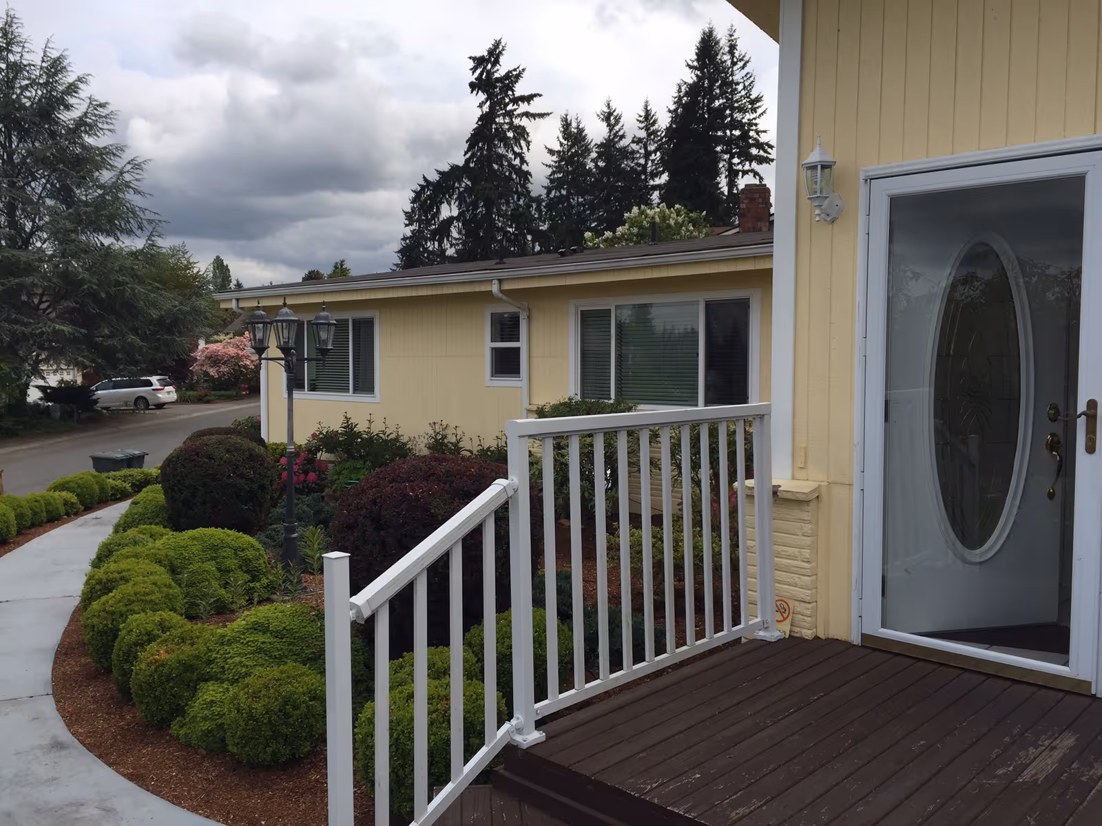 View of the front entrance of a yellow single-story building with a white railing along the wooden porch steps. There are well-maintained bushes and shrubs along a curved concrete walkway leading to the entrance. A street lamp with three lanterns is visible near the walkway, and tall evergreen trees are in the background under a cloudy sky.