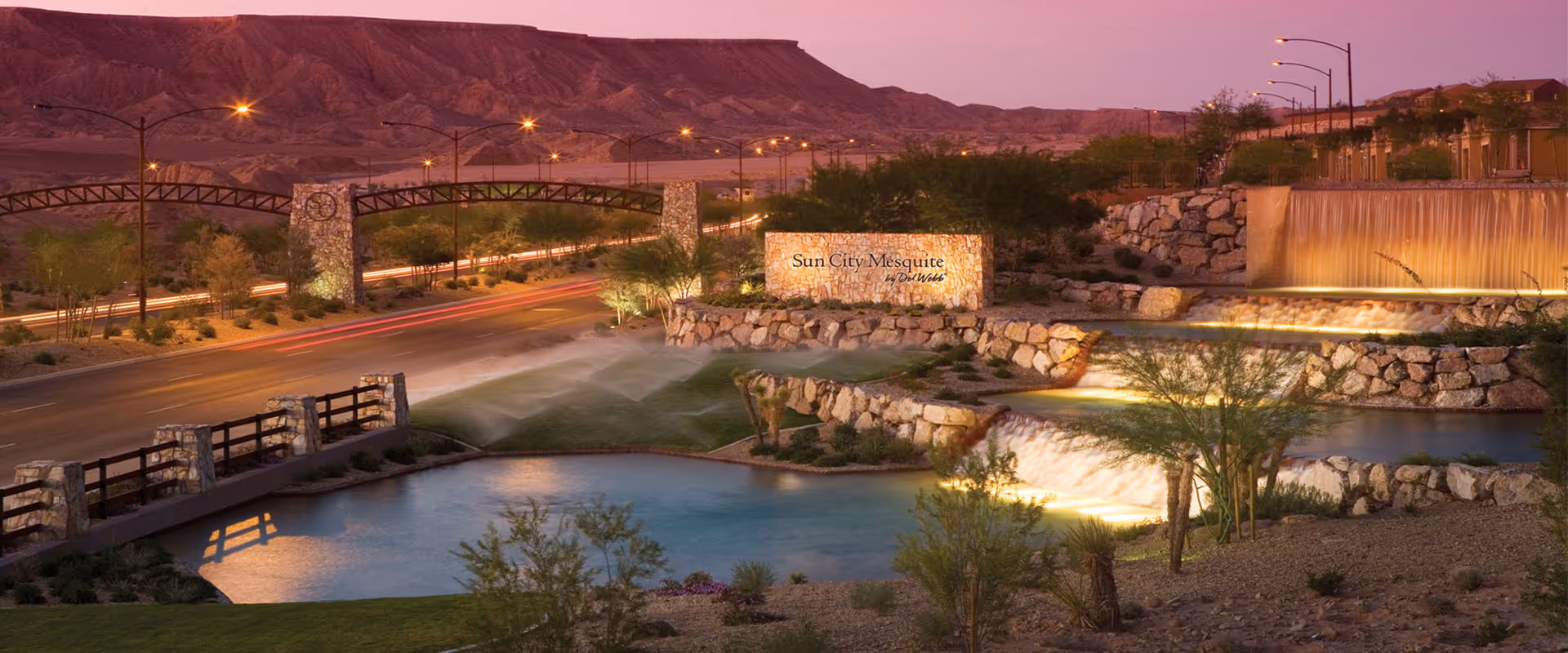 Evening view of the entrance to Sun City Mesquite by Del Webb, featuring a landscaped area with water features including waterfalls and a pond, stone signage with the community name, street lamps, and a mountainous backdrop under a purple sky.