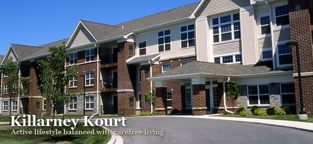 Exterior view of a multi-story brick and siding senior living facility with multiple windows, a covered entrance, and a curved driveway surrounded by small trees and landscaping under a clear blue sky.