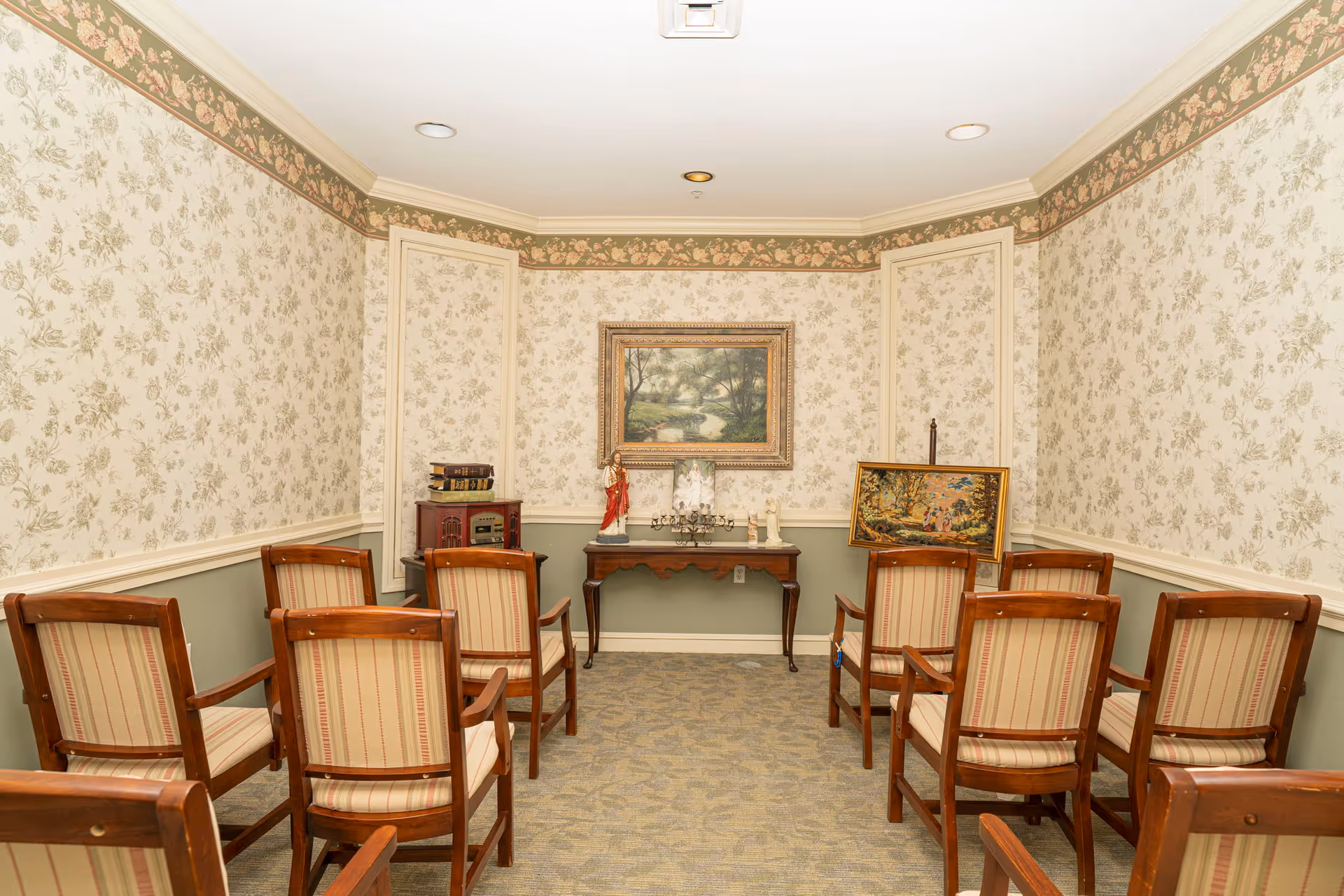A small room with floral wallpaper and a green lower wall border, furnished with two rows of wooden chairs with striped cushions facing a wooden table against the far wall. On the table are decorative items including a statue and a candelabra, with framed paintings hanging and leaning against the wall behind it.