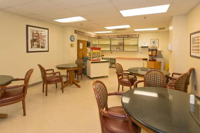 A well-lit room with several round tables and chairs arranged around them. The chairs have wooden frames with cushioned seats and backs in a plaid pattern. The walls are light-colored and decorated with framed artwork. At the back of the room, there is a counter area with cabinets and a popcorn machine on a small cart. The ceiling has recessed fluorescent lighting.