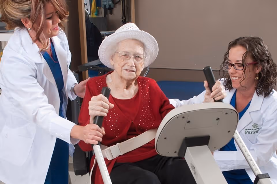 An elderly woman wearing a white hat and red sweater is seated on a stationary exercise bike, assisted by two female healthcare professionals in white coats. They are helping her hold the bike handles and encouraging her during the exercise session inside a rehabilitation facility.