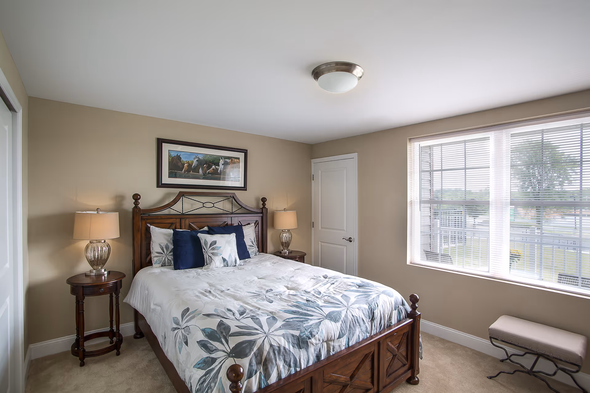 A bedroom with a wooden bed frame and a floral patterned bedspread. There are two bedside tables with lamps on each side of the bed. A framed picture of horses hangs above the bed. A window with blinds lets in natural light, and a small bench is placed under the window. The walls are painted beige and the carpet is light-colored.