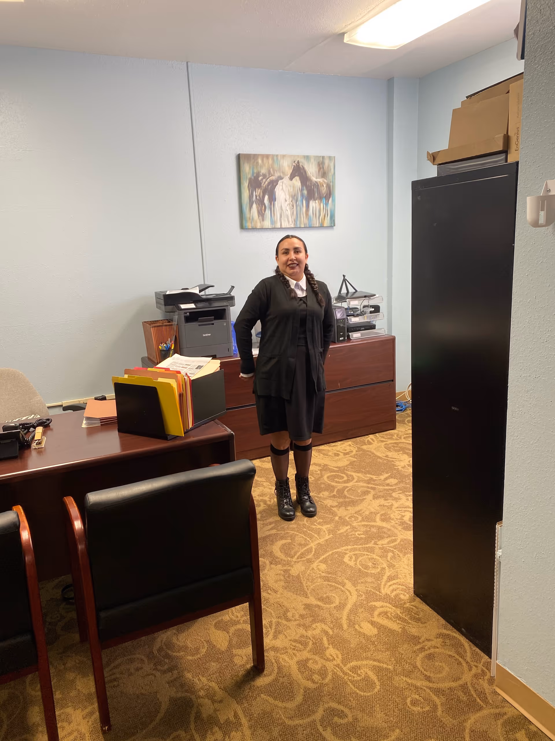 A woman standing in an office room with light blue walls and patterned carpet. The room contains a wooden desk with office supplies and file folders, two black chairs with wooden armrests, a printer on a cabinet, and a painting of horses on the wall.