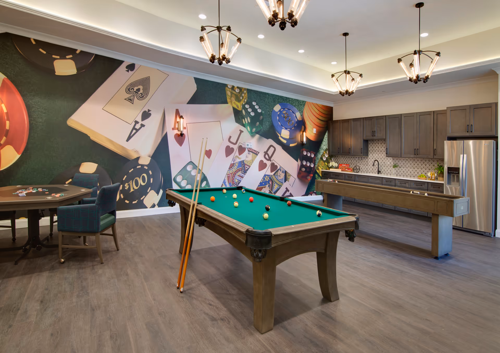 A recreational room featuring a pool table with balls arranged on the green felt surface and two pool cues resting against it. To the left, there is a card table with poker chips and two blue plaid chairs. The back wall has a large mural depicting playing cards, poker chips, and dice. On the right side of the room, there is a kitchenette area with dark wood cabinets, a stainless steel refrigerator, a sink, and a shuffleboard table in front of it. The room is well-lit with modern ceiling lights and wall sconces.