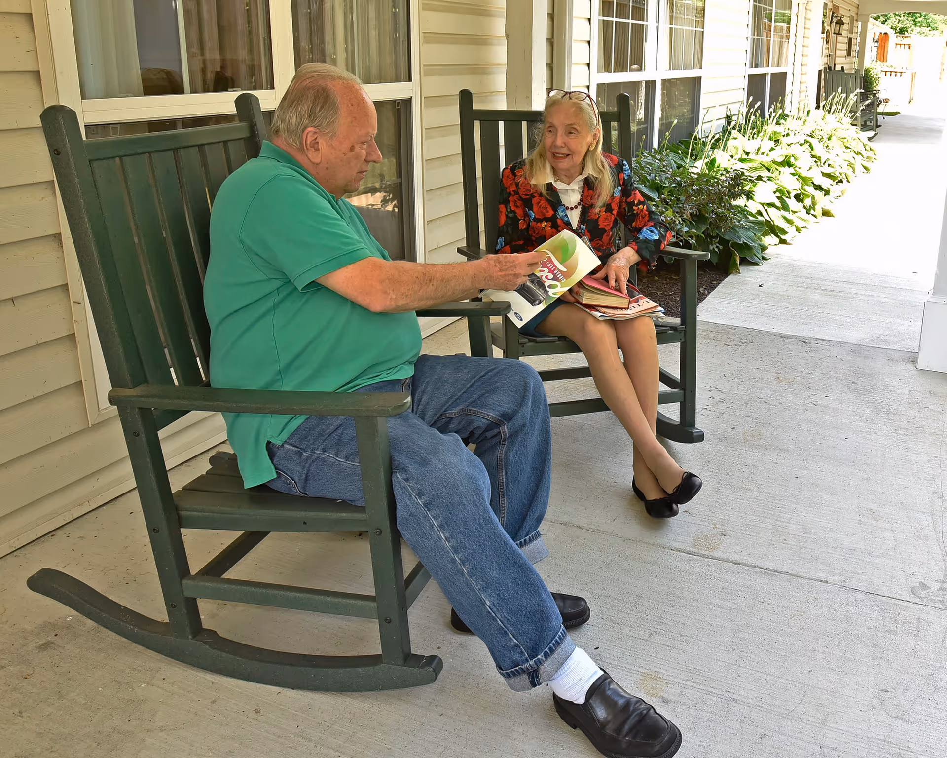 An elderly man and woman sitting on green rocking chairs on a covered porch. The man is wearing a green polo shirt and jeans, holding a magazine, while the woman, dressed in a floral jacket and black shoes, is smiling and holding a book. There are plants along the side of the porch and windows behind them.