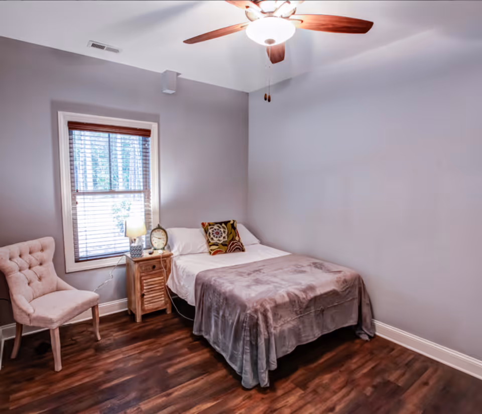 A small bedroom with a single bed covered with a gray blanket and a decorative pillow. Next to the bed is a wooden nightstand with a lamp and a clock. There is a beige upholstered chair near a window with wooden blinds. The room has wooden flooring and a ceiling fan with a light.
