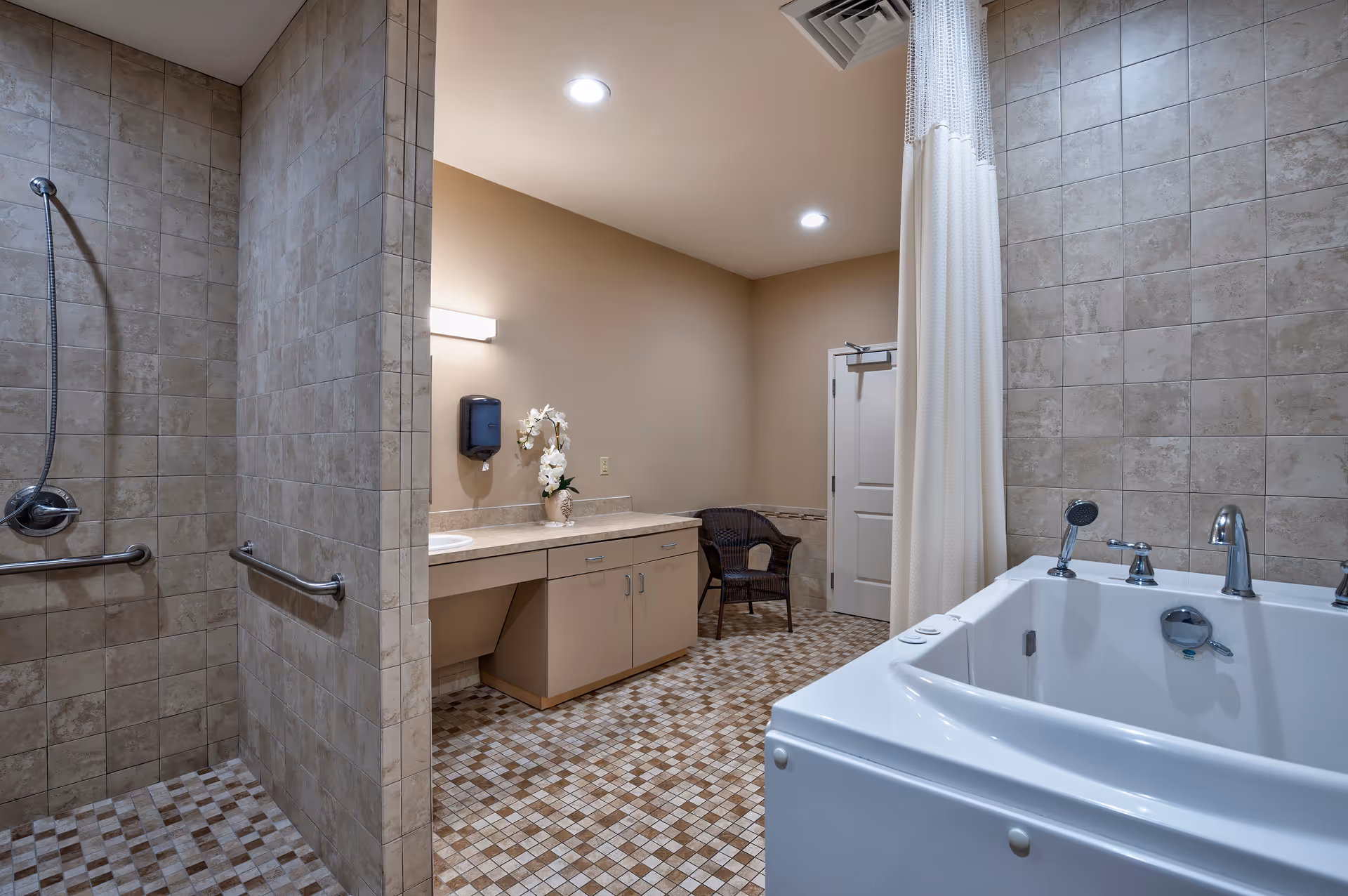 A spacious bathroom with beige tiled walls and mosaic tiled floor. The room features a walk-in shower with grab bars, a large white bathtub with multiple faucets, a countertop with a sink, a soap dispenser mounted on the wall, a vase with white flowers, a wicker chair, and a closed white door in the background.