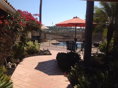 Sunlit outdoor patio with tiled walkway, plants, tables, umbrellas, and a pool area.