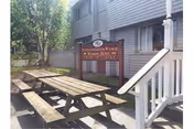 Outdoor area of Fiddler's Green Manor Home featuring wooden picnic tables and benches on a wooden deck, with a tree providing shade and the facility building in the background displaying a sign with the facility's name.