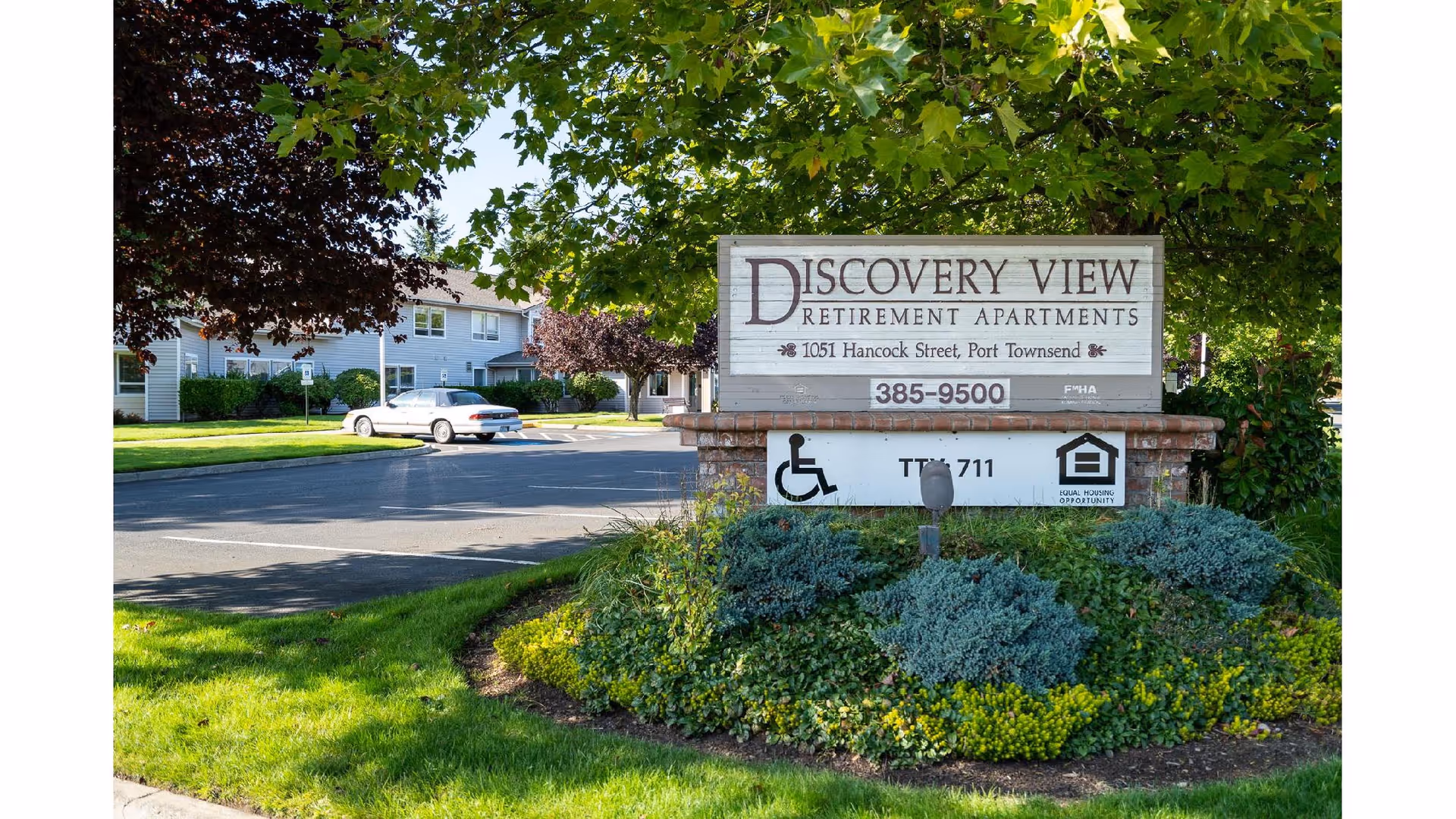 Entrance sign for Discovery View Retirement Apartments located at 1051 Hancock Street, Port Townsend, surrounded by green shrubs and trees with a residential building and parked car in the background.