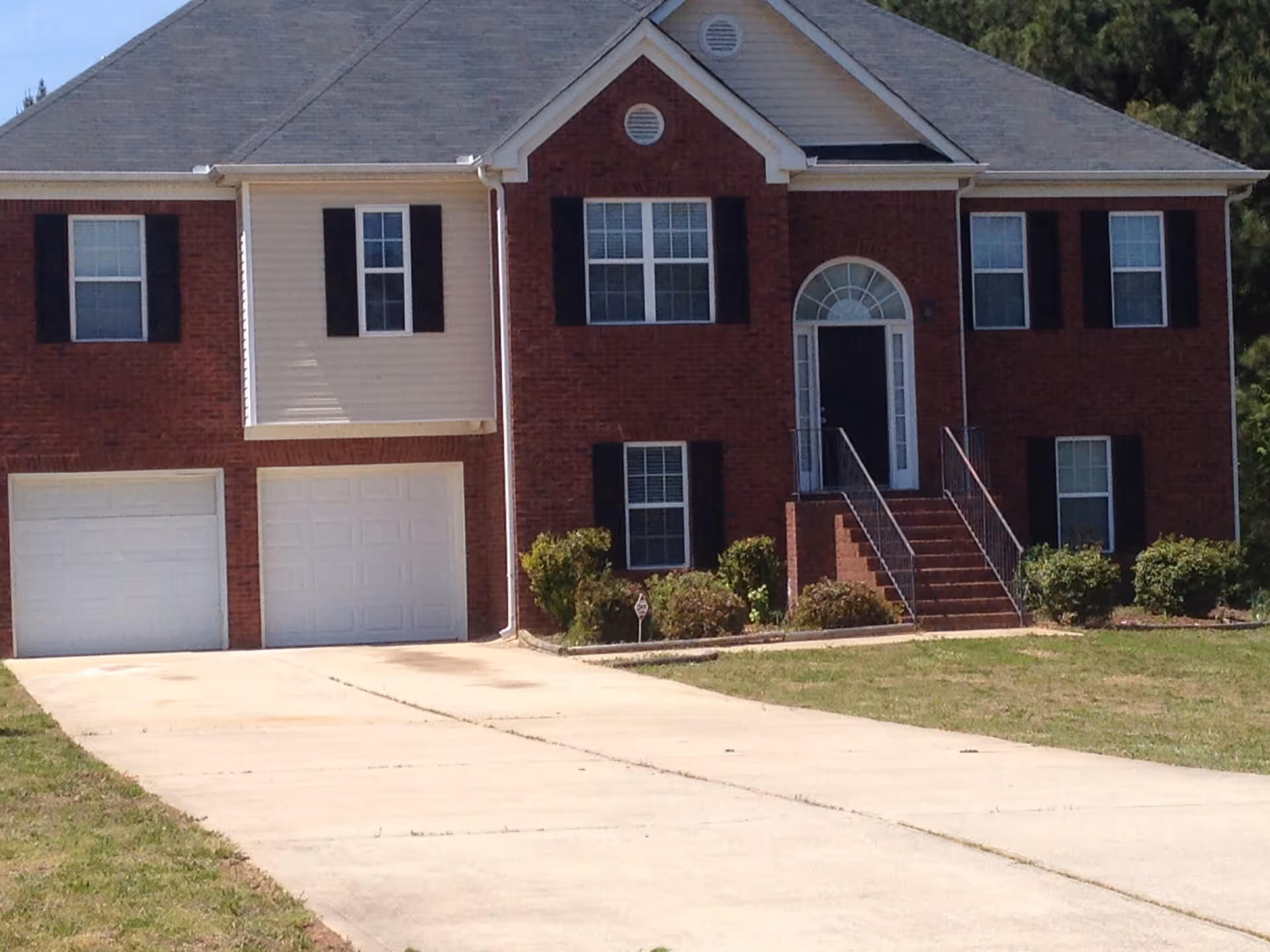Front exterior view of a two-story brick house with a gray roof, black shutters, a double garage, and a concrete driveway leading up to the entrance with stairs and a decorative arched window above the door. There are bushes and a lawn in front of the house.