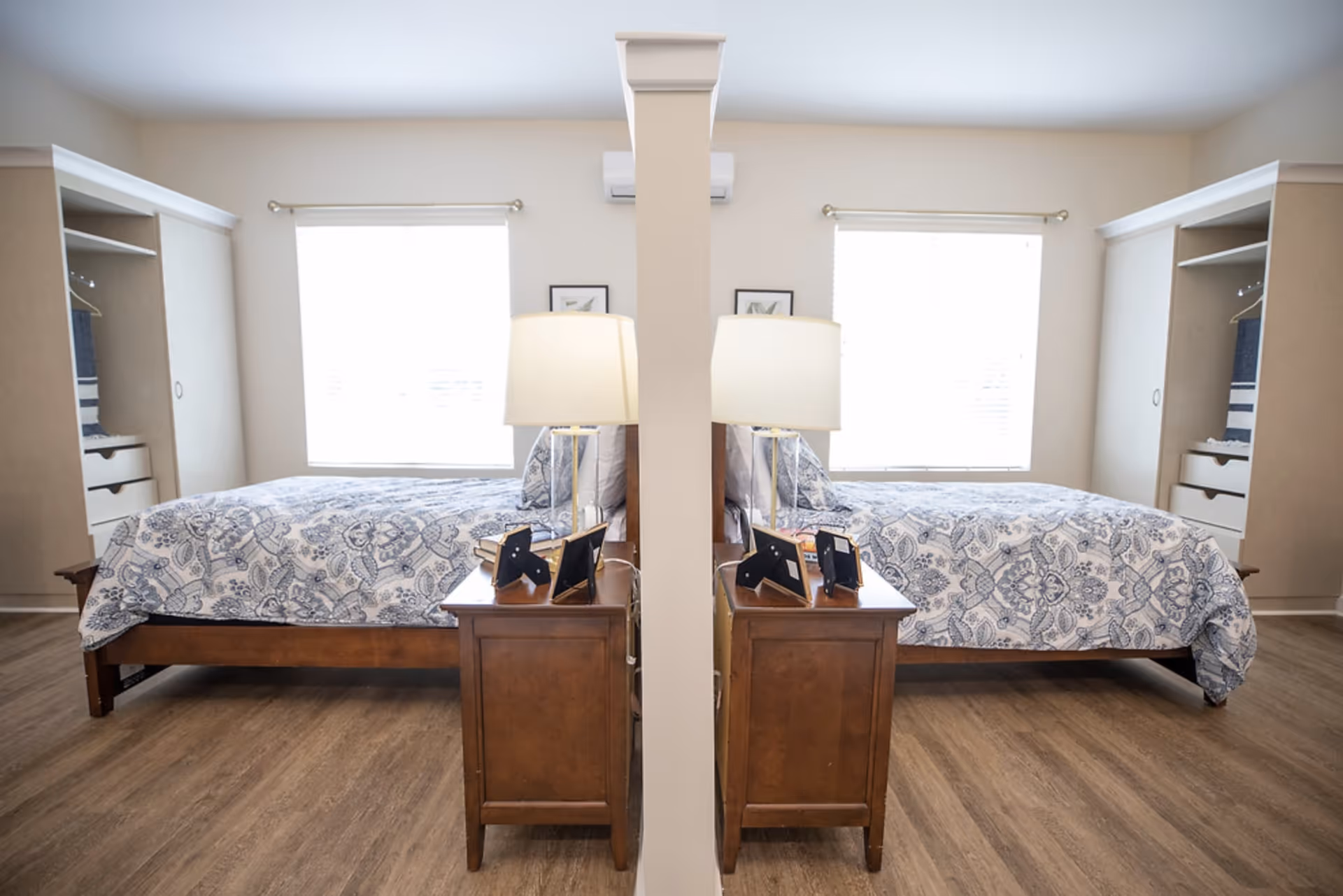 A bright bedroom with a wooden bed featuring a blue and white patterned bedspread. There are two windows with white blinds on the back wall, and a wooden nightstand with a lamp and picture frames is positioned next to the bed. The room has light-colored walls and wood flooring, with built-in closets on either side of the bed.