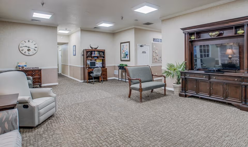 A cozy living room area in a senior living facility with beige walls and carpeted floor. The room features a large wooden entertainment center with a flat-screen TV, a green upholstered loveseat, a beige recliner chair, a wooden desk with a computer and chair, a small side table, a wall clock, framed artwork, and a potted plant.