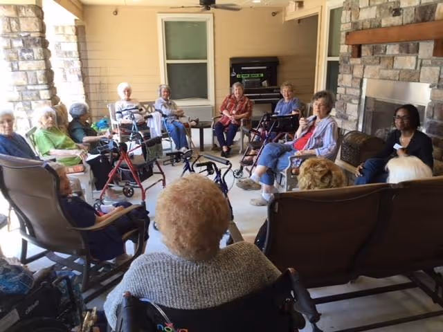 A group of elderly individuals sitting in a circle on a covered outdoor patio at Landon Ridge Sugar Land Independent Living. Some are seated in wheelchairs and others in chairs, engaging in conversation. The patio features stone pillars and a stone fireplace.