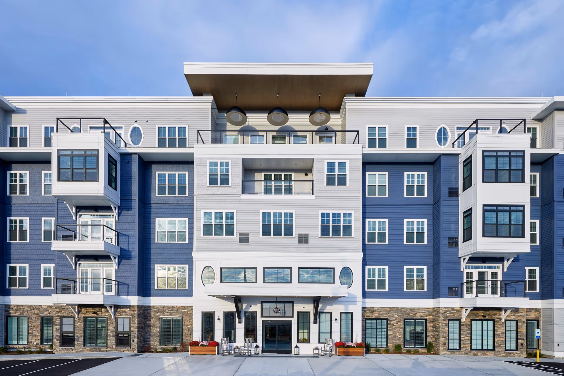 Front exterior view of a modern multi-story senior living facility with blue and white siding, large windows, balconies, and a covered entrance with seating and planters.